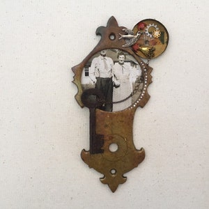 May include: A vintage key-shaped frame with a black and white photo of a couple standing in front of a house. The frame is decorated with a small bird, gears, and beads.