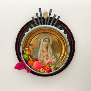 May include: A religious icon featuring a vintage image of a saint, surrounded by a gold-toned frame and a decorative arrangement of faux fruits and flowers. The artwork is set within a circular, dark brown frame with a light blue border and metal accents.