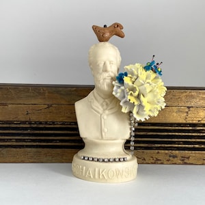 May include: A white porcelain bust of a man with a beard, wearing a dark jacket. The bust is decorated with a small brown bird, a yellow flower, and a silver chain. The base of the bust is inscribed with the name "Tchaikovsky".
