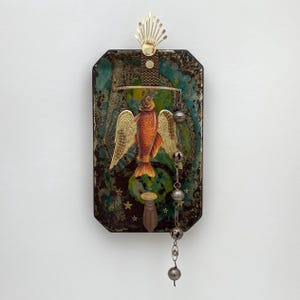 May include: A rectangular wall plaque featuring a dark, mottled background with green and gold accents. A central orange fish with gold wings is mounted on the surface. A gold metal crest sits at the top, and a chain of small silver bells hangs down.