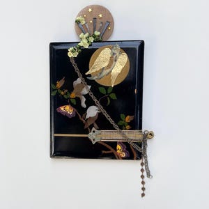 May include: A decorative black rectangular wall art piece with a gold angel, butterflies, and floral accents. A wooden circle with stars and flowers is above the rectangle. A silver chain and decorative bar are attached.