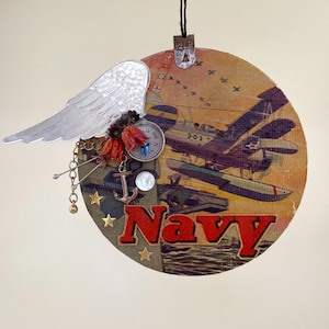 May include: A round wooden ornament with a vintage image of a seaplane and the word "Navy" in red letters. The ornament is decorated with a silver angel wing, a compass, an anchor, and gold stars.