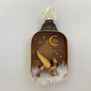 May include: A decorative, rectangular box featuring a brown and gold celestial scene with a crescent moon and stars. A small dog with gold wings rests on a bed of white feathers. The box is topped with a gold and bronze decorative element.