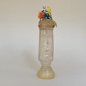 May include: A clear glass bottle with a sculpted face and a gold crown. The bottle is decorated with a cluster of colorful flowers.