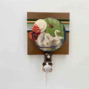 May include: A square brown wooden plaque featuring a circular print of two white swans on water, accented with a miniature straw hat, orange fabric flowers, a striped ribbon, and a hanging bundle of twigs with a white leaf-shaped charm.