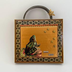 May include: A decorative box with a gold background featuring a frog illustration, gold stars, and a metal handle. The box has a decorative border with green and red accents, and a gold dragonfly charm.