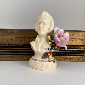 May include: A white ceramic bust of Mozart with a pink flower and colorful beads on the shoulder. The bust is on a white base with the word "MOZART" written on it.