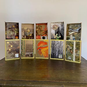 May include: A collection of five decorative matchboxes, each with unique artwork and embellishments. The boxes feature various themes, including a castle, a rooster, and a vintage airplane. The boxes are displayed on a dark wooden surface.
