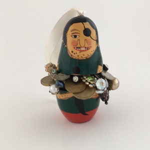May include: A hand-painted wooden ornament shaped like a Russian nesting doll, with a green and red design. The doll's face is painted with a black eye patch and a mischievous grin. The doll is adorned with a variety of colorful jewels and beads.
