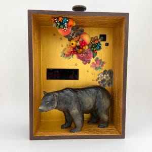 May include: A decorative wooden box with a gold interior featuring a black bear figurine. The box is adorned with floral and fruit decals. The bear is dark gray and stands in front of a rectangular black shape.