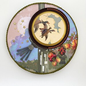 May include: A round decorative piece with a framed illustration of ducks and a turtle flying. The piece has a pink and blue background with gold stars and a green trim. There are also illustrations of fruit.