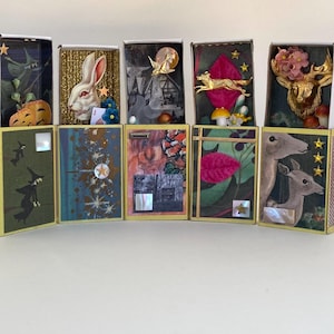 May include: A selection of decorative matchboxes with diverse designs. The boxes are adorned with a rabbit, a deer, and a witch. The color scheme includes gold, green, and blue, with white and pink highlights. The matchboxes are arranged in a line.