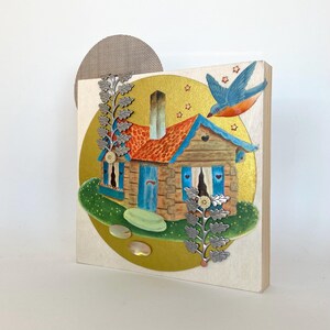 May include: A small, blue and brown cottage with a red roof sits on a green lawn with a yellow moon behind it. The cottage has a blue door and two windows with blue shutters. There are two silver leaf branches on either side of the cottage. A blue bird with orange accents flies above the cottage.