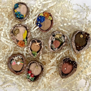 May include: A collection of decorative walnut shell art pieces. Each shell features a sculpted face with various floral and nature-inspired embellishments in shades of blue, orange, and gold. The shells are arranged on a bed of white paper filler.