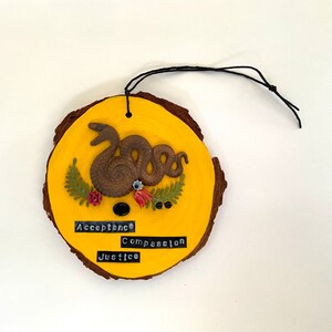Peut inclure: Une décoration ronde en bois avec un fond jaune et un motif de serpent brun. La décoration porte les mots "Acceptance", "Compassion" et "Justice".
