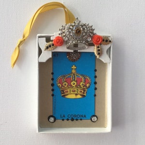 May include: A handmade ornament featuring a vintage Loteria card with the image of a crown, "La Corona", and the number 47. The card is framed in a white box with a jeweled crown and red flowers on top.