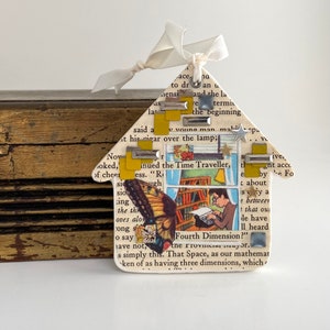 May include: A house-shaped ornament made from a book page with a butterfly and a typewriter image. The ornament is decorated with yellow and silver accents and a white ribbon.