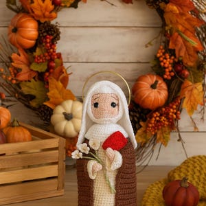 Puede incluir: Muñeca de ganchillo hecha a mano de una santa, con velo blanco y túnica marrón, sosteniendo flores y un corazón rojo. La muñeca está colocada frente a una corona de otoño, calabazas y una caja de madera, evocando un tema otoñal.