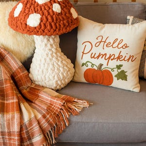 May include: A cozy autumn-themed scene featuring a large mushroom-shaped pillow with white spots, a pillow with "Hello Pumpkin" text and pumpkin graphic, and a plaid throw blanket in orange and cream colors. Decorative pumpkins complete the fall decor.