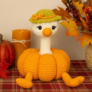 May include: A handcrafted crochet goose with a green hat, orange pumpkin body and feet. The goose is on a plaid surface, beside a pumpkin, a candle, and a vase of autumn leaves. The theme is autumn and harvest.