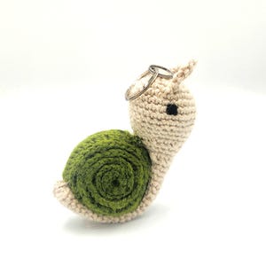 Puede incluir: Un llavero de caracol tejido a ganchillo con caparazón verde y cuerpo beige. El caracol tiene un ojo negro y un llavero plateado sujeto a la cabeza. El artículo hecho a mano está sobre un fondo blanco.