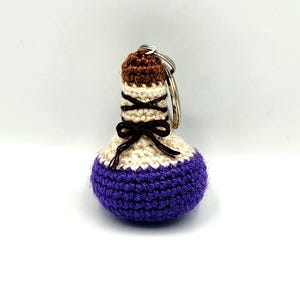 Puede incluir: Un llavero de ganchillo hecho a mano con forma de botella de poción. La botella presenta una base morada, una sección media de color crema y una parte superior marrón. Un pequeño lazo marrón está atado alrededor de la sección crema. Un llavero plateado está unido a la parte superior.