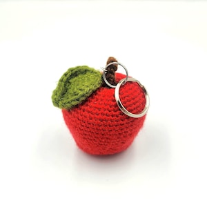 May include: A red crocheted apple keychain with a green leaf and a silver key ring. The apple is a vibrant red color, and the leaf is a contrasting green. The keychain is a handmade item.