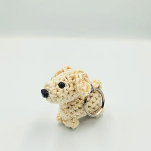 May include: A light beige crocheted dog keyring. The dog has black eyes and a black nose. A silver-coloured metal ring is attached to the dog's back. The keyring is a handmade item.