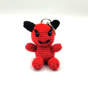 Puede incluir: Un llavero de diablo rojo tejido a mano con cuernos negros y ojos negros. El diablo tiene una cara sonriente y brazos y piernas pequeños. El llavero está sujeto a un anillo de metal plateado.