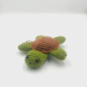May include: A handmade crochet turtle keychain. The turtle features a brown shell, green body, and small black eyes. The keychain is made of yarn and includes a silver-colored metal ring.