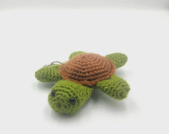 May include: A handmade crochet turtle keychain. The turtle features a brown shell, green body, and small black eyes. The keychain is made of yarn and includes a silver-colored metal ring.