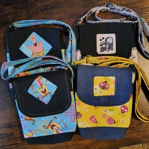 May include: Four small crossbody bags with adjustable straps. Two bags feature Spongebob Squarepants characters on blue fabric. One bag has a Jack and Sally patch, and the other has a ladybug design on yellow fabric. All bags have black flaps.