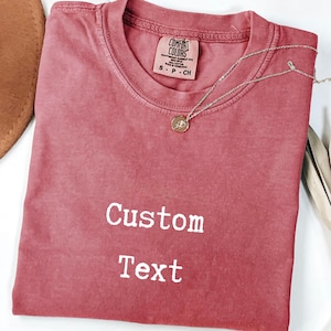 May include: A dusty rose-colored t-shirt with the words "Custom Text" in white. A silver necklace with a pendant is draped across the shirt. The shirt has a tag that reads "Comfort Colors". A brown hat is partially visible.