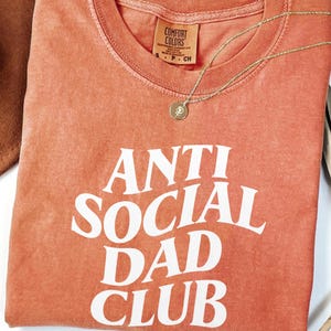 Puede incluir: Una camiseta de color coral con las palabras "ANTI SOCIAL DAD CLUB" en letras blancas. Un collar dorado con un colgante descansa sobre la camiseta. Un sombrero marr&oacute;n es parcialmente visible en la esquina superior izquierda.