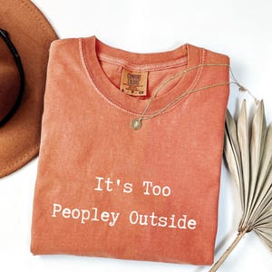 May include: A coral-colored t-shirt with the text "It's Too Peopley Outside" in white. A gold necklace with a pendant rests on the shirt. A brown hat and dried palm leaves are in the background. The shirt is a Comfort Colors brand.