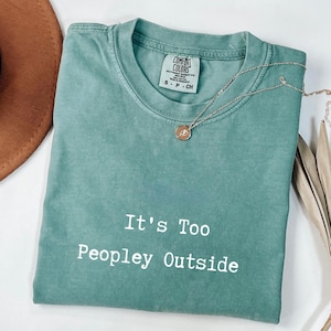 May include: A seafoam green t-shirt with the text "It's Too Peopley Outside" in white. A gold-colored necklace with a pendant is draped across the shirt. The shirt is folded, and a brown hat is visible in the background.
