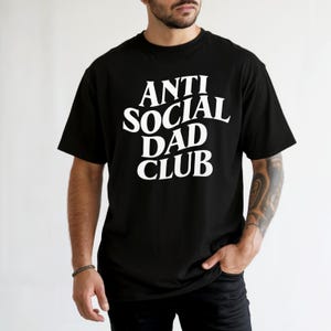 Puede incluir: Camiseta negra con el texto blanco "ANTI SOCIAL DAD CLUB" en una fuente en negrita sin serifa. La camiseta es de estilo cuello redondo cl&aacute;sico, y el texto est&aacute; centrado en el pecho.