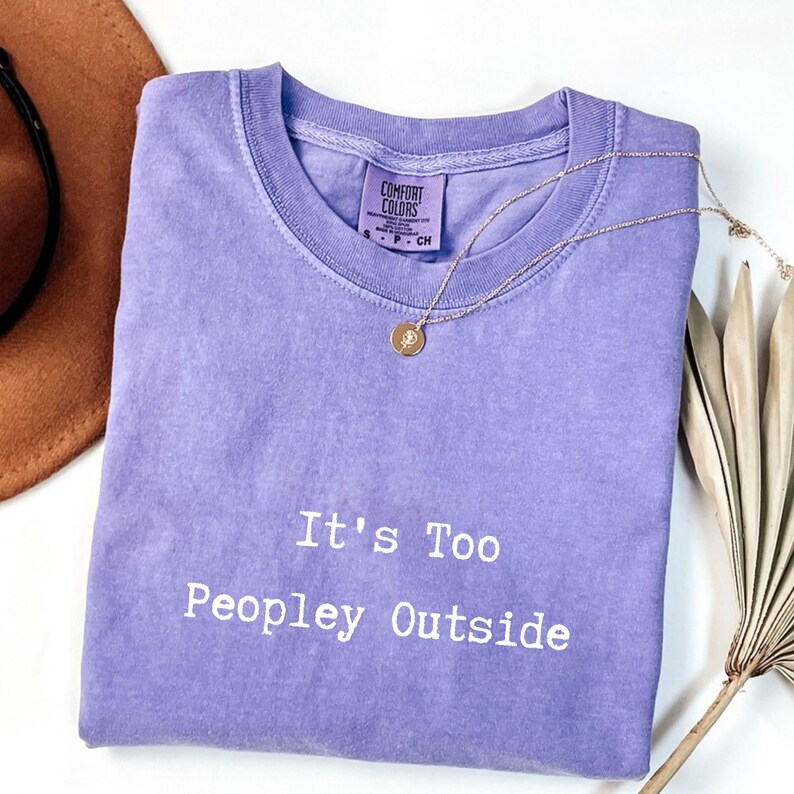 May include: A lavender Comfort Colors t-shirt with the phrase "It's Too Peopley Outside" in white text. A gold necklace with a pendant rests on the shirt. A brown hat is visible in the upper left corner.