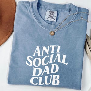 Puede incluir: Una camiseta azul con la frase "ANTI SOCIAL DAD CLUB" en blanco. Un collar dorado con colgante descansa sobre la camiseta. La camiseta est&aacute; doblada y tiene una etiqueta Comfort Colors. Talla S.