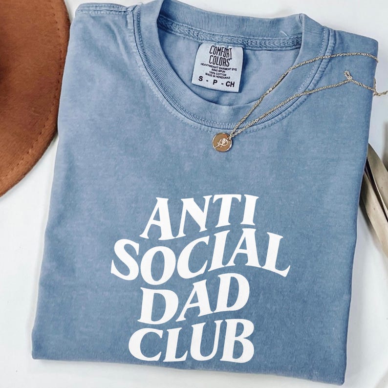 Puede incluir: Una camiseta azul con la frase "ANTI SOCIAL DAD CLUB" en blanco. Un collar dorado con colgante descansa sobre la camiseta. La camiseta est&aacute; doblada y tiene una etiqueta Comfort Colors. Talla S.