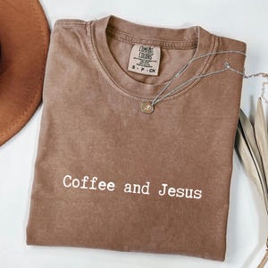 May include: A brown Comfort Colors t-shirt with the text "Coffee and Jesus" in white. A silver necklace with a pendant is draped across the shirt. A brown hat is visible in the upper left corner.