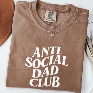 Puede incluir: Una camiseta marr&oacute;n con el texto "ANTI SOCIAL DAD CLUB" en letras blancas. La camiseta est&aacute; doblada y tiene una etiqueta Comfort Colors. Un collar plateado con un colgante est&aacute; encima de la camiseta.