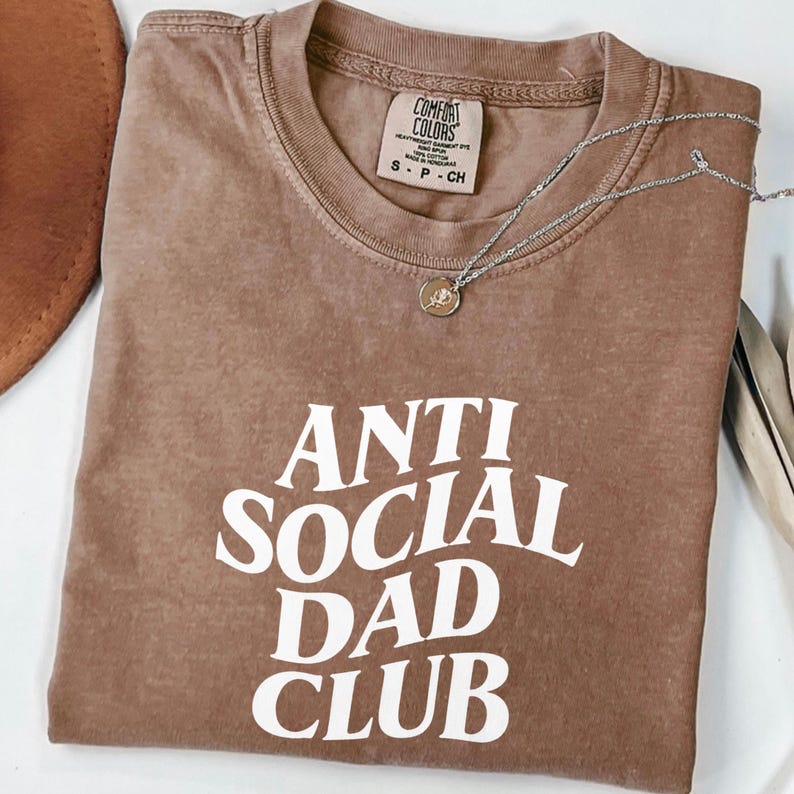 Puede incluir: Una camiseta marr&oacute;n con el texto "ANTI SOCIAL DAD CLUB" en letras blancas. La camiseta est&aacute; doblada y tiene una etiqueta Comfort Colors. Un collar plateado con un colgante est&aacute; encima de la camiseta.