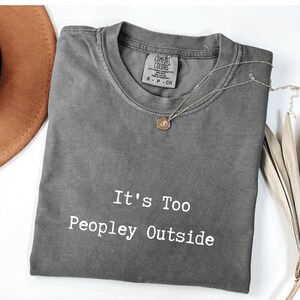 May include: A gray Comfort Colors t-shirt with the text "It's Too Peopley Outside" in white. A silver necklace with a pendant and a brown hat are also visible. The shirt is folded and has a vintage look.
