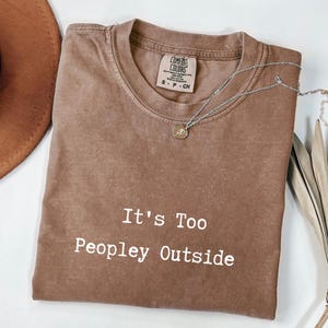 May include: Brown Comfort Colors t-shirt with the text "It's Too Peopley Outside" in white. A silver necklace with a small pendant rests on the shirt. The shirt is folded and displayed with a brown hat and dried leaves.