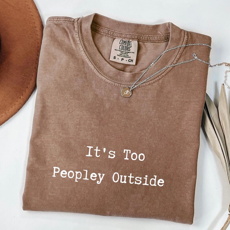 May include: Brown Comfort Colors t-shirt with the text "It's Too Peopley Outside" in white. A silver necklace with a small pendant rests on the shirt. The shirt is folded and displayed with a brown hat and dried leaves.