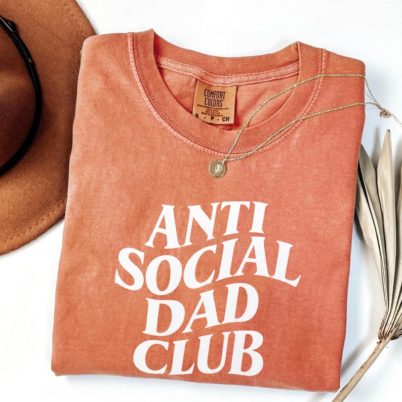 Puede incluir: Una camiseta de color coral con las palabras "ANTI SOCIAL DAD CLUB" en letras blancas. Un collar dorado con un colgante descansa sobre la camiseta. Un sombrero marr&oacute;n es parcialmente visible en la esquina superior izquierda.