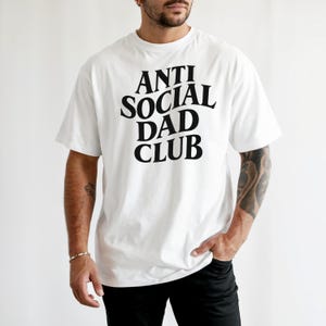 Puede incluir: Camiseta blanca con el texto "ANTI SOCIAL DAD CLUB" en una fuente negra y audaz. La camiseta de manga corta parece estar hecha de un material suave. La persona lleva pantalones negros.