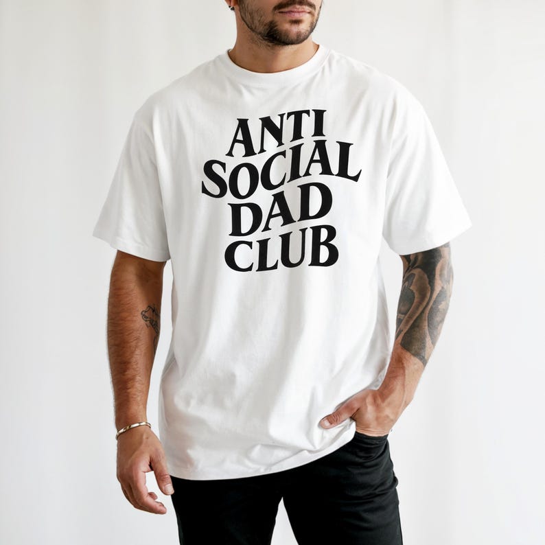 Puede incluir: Camiseta blanca con el texto "ANTI SOCIAL DAD CLUB" en una fuente negra y audaz. La camiseta de manga corta parece estar hecha de un material suave. La persona lleva pantalones negros.