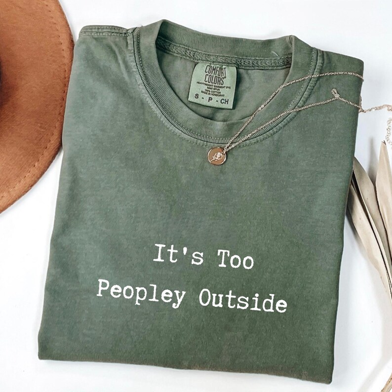 May include: An olive green t-shirt with the text "It's Too Peopley Outside" in white. A gold necklace with a pendant rests on the shirt. A brown hat is partially visible in the upper left corner.